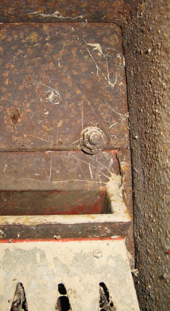 Inside a modified wall box, looking up into the top corner of the box with replacement aperture below. One of the four studs holding the larger aperture plate in place can be seen. Below the aperture are the 'dragons teeth' designed to prevent theft of mail from a post box